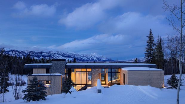 A contemporary single-storey residence set in a snowy alpine landscape, viewed from outside at dusk. The home has a low, mostly flat roofline, warm glowing floor-to-ceiling glazing, pale timber or rendered wall surfaces, and a rounded projecting wing. Snow-covered ground, coniferous trees, and a dramatic mountain range surround the building.