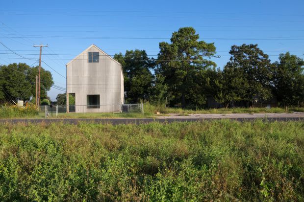 A small, simple gable-roofed building viewed from across an overgrown roadside verge. The structure has pale vertical corrugated cladding, a central dark window on the front elevation, a smaller upper-level window within the gable, and a side door. Chain-link fencing, utility poles and overhead wires surround the site, with mature trees behind it. The scene has a utilitarian rural or semi-rural character under clear blue skies.