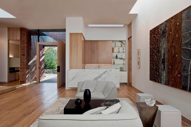 A refined open-plan living area with warm timber flooring and joinery, a white marble-clad island or bar, glass display shelving, and a sculptural contemporary artwork. The room connects to an entry or circulation area through a glazed timber door, with exposed brickwork and skylights adding architectural character. Neutral upholstered seating, dark occasional furniture, and recessed ceiling lighting create a calm, polished interior.