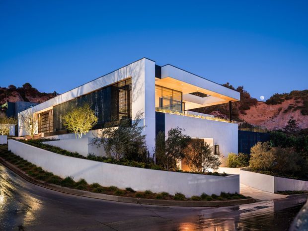 Twilight exterior view of a large contemporary multi-level residence on a sloping urban site. The home features crisp white rendered volumes, extensive floor-to-ceiling glazing, dark vertical screening, broad cantilevered roof planes, warm concealed lighting, landscaped terraces, and a curved boundary wall along a wet street. Rocky hills and mature vegetation form the background.