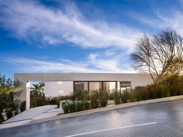 A low, single-storey modern residence viewed from across a sloping paved street. The home has a long, minimalist white rendered facade with a broad flat roofline, recessed entry, and dark-framed floor-to-ceiling glazing. Layered ornamental grasses and other drought-tolerant landscaping soften the facade, while a concrete path leads toward the entrance. The image is captured in bright daylight with a blue, lightly clouded sky.