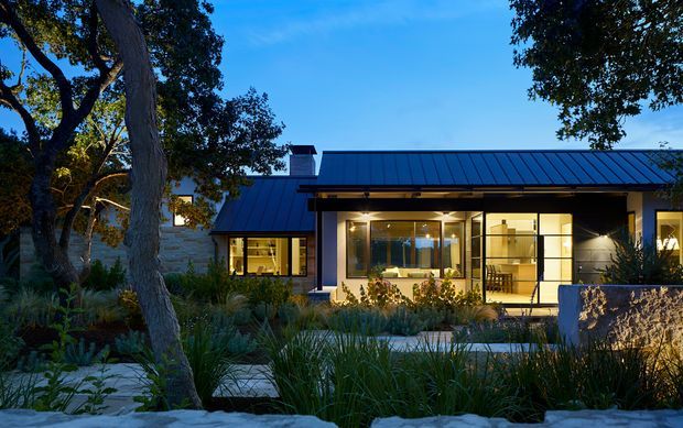Twilight exterior view of a low, single-storey contemporary house with a long dark standing-seam metal roof, extensive illuminated glazing, pale masonry walls, and a landscaped foreground with native-style planting and a paved entry path. Mature trees frame the facade.