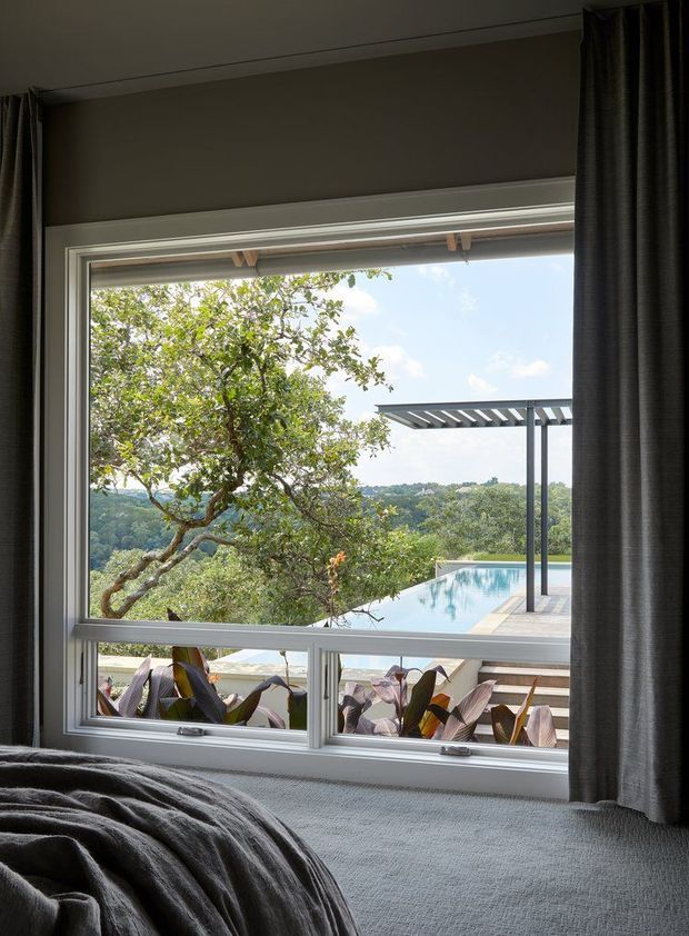 A moody bedroom interior viewed toward a large framed window, with a textured grey bed and dark curtains in the foreground. Through the glazing is a leafy landscaped setting with a swimming pool, tiled terrace, and modern pergola, creating a strong indoor-outdoor connection. The room has a restrained, contemporary feel with neutral grey finishes and abundant natural light beyond the window.
