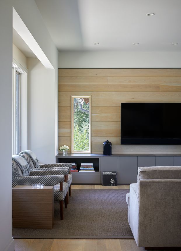 A refined living room with a large wall-mounted television set into a horizontal light timber feature wall, paired with dark grey built-in cabinetry and open shelving below. The room includes patterned armchairs, a pale upholstered sofa, a timber side table, a broad neutral rug, recessed ceiling lights, and a narrow vertical window providing greenery views. The overall design is warm, contemporary, and restrained.