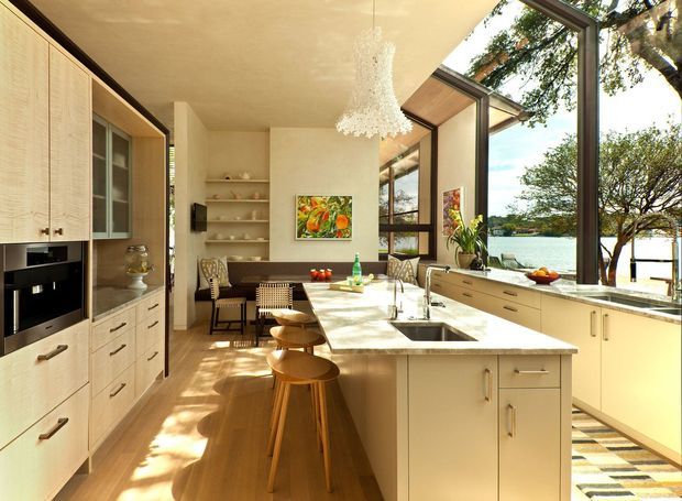 Bright open-plan kitchen and dining area with extensive floor-to-ceiling glazing overlooking a waterfront landscape. The room features light timber cabinetry, a large pale island with integrated sink and seating, open shelving, warm timber flooring, and a sculptural white pendant light. Natural daylight creates a warm, airy atmosphere.