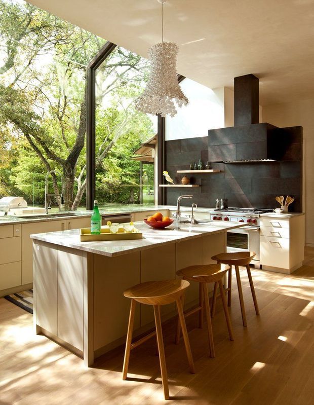 A bright, spacious contemporary kitchen with a large central island and four timber bar stools. Floor-to-ceiling glazing opens the room to leafy greenery outside, while a dark stone or slate feature wall frames the cooktop and rangehood. Light timber flooring, pale cabinetry, a stone island benchtop, open timber shelving, and a sculptural clustered pendant create a warm, refined interior.