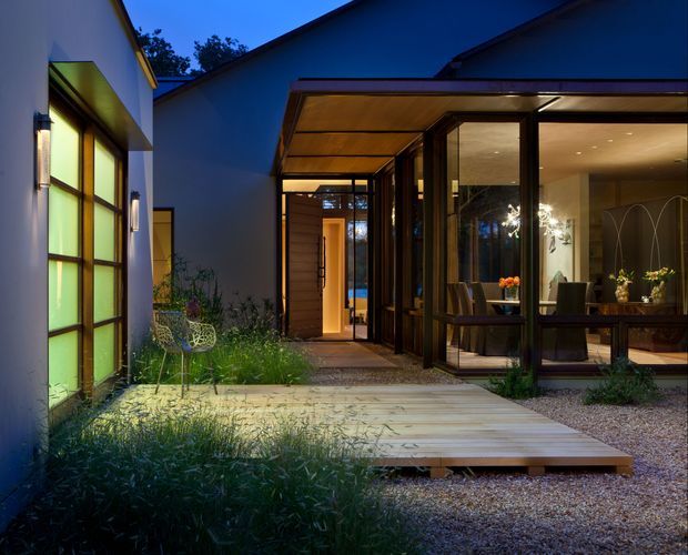 Twilight view of a contemporary residential courtyard and entry, with dark-framed floor-to-ceiling glazing, warm timber soffits and decking, illuminated interior dining and living spaces, and landscaped planting around a gravel ground plane.