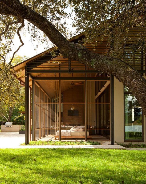 Exterior view of a warm timber-clad house set among mature trees and lawn. The facade features a steeply sloping roof, extensive floor-to-ceiling glazed screens with dark slender frames, a covered timber-framed entry or sunroom, and a bedroom visible through the glazing. A large tree crosses the foreground, while a paved outdoor area and built-in barbecue are visible to the left.