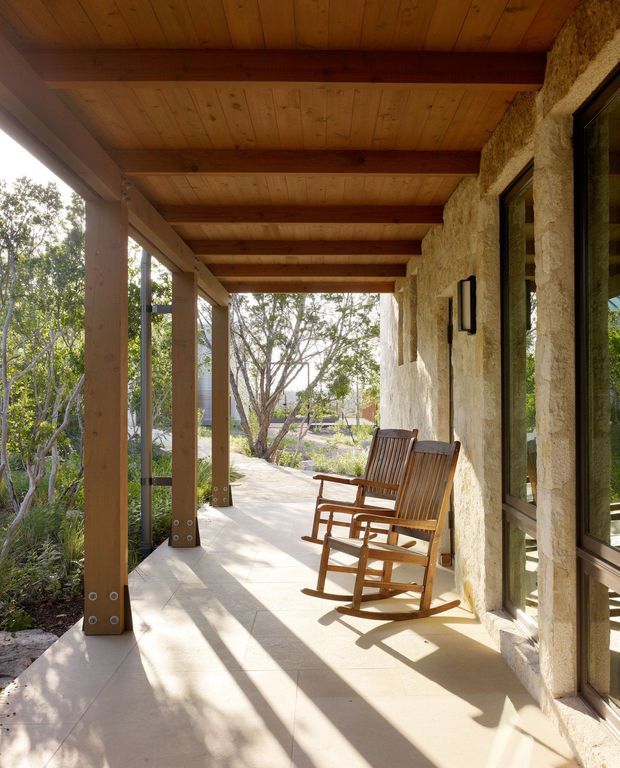 A sunlit covered outdoor veranda runs along a rustic residence, with substantial timber posts and beams supporting a timber-lined ceiling. The wall is finished in rough natural stone or textured masonry, with dark-framed glazing and two wooden rocking chairs facing a landscaped garden. Long shadows, warm timber tones, and natural materials create a relaxed connection between the house and garden.