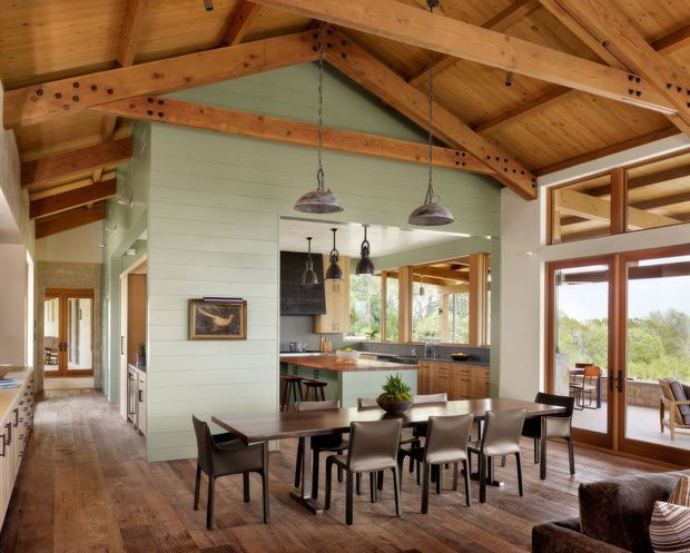Open-plan dining and kitchen interior with a long dark timber dining table and upholstered dining chairs in the foreground. The room features exposed timber post-and-beam roof framing, a pitched timber-lined ceiling, pale green horizontal wall cladding, timber flooring and cabinetry, pendant lights, and extensive timber-framed glazing opening toward an outdoor terrace. A hallway and living area extend beyond the dining space.