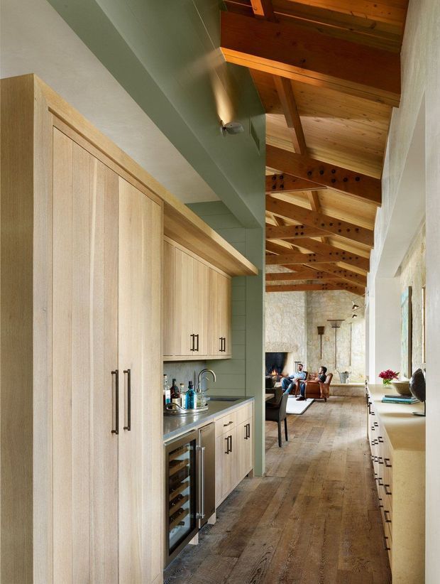 A warm, elongated open-plan interior showing a compact built-in kitchen and bar joinery in the foreground, opening into a rustic living area beyond. The kitchen features light timber cabinetry with dark linear handles, a stainless-steel benchtop, integrated underbench wine refrigerator, sink and pale green tiled backsplash. The living space has a stone feature wall, timber flooring, leather seating and a high exposed timber-beam ceiling with visible rafters. A long pale built-in storage or seating unit runs along the right wall.