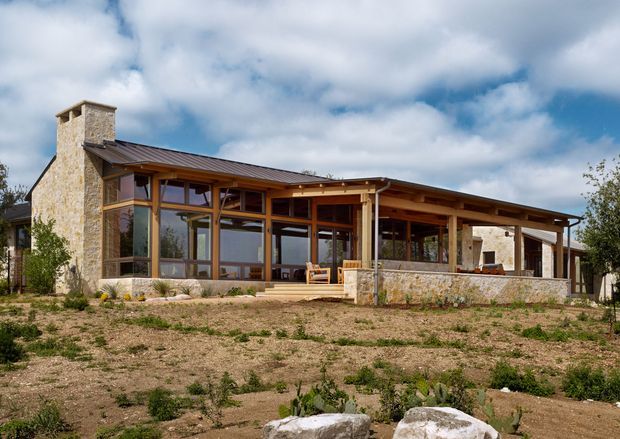 Exterior view of a low-rise, lodge-like residence set in a sparse landscaped site. The building features a broad low-pitched metal roof, extensive timber-framed glazing, exposed timber structural elements, and substantial natural-stone chimney and wall construction. A covered terrace/veranda extends across the front, with stone retaining walls and steps.
