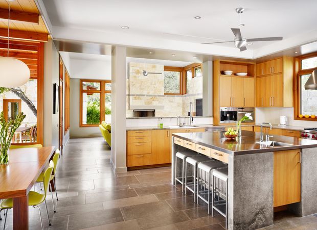 Bright, spacious open-plan kitchen with extensive warm timber cabinetry, a large stone island with integrated seating, stainless-steel appliances, and a pale stone feature wall surrounding the cooking area. The kitchen opens toward a dining area and living space, with abundant timber-framed glazing, white walls, recessed lighting, and a ceiling fan. The overall design is clean, warm, and contemporary with natural materials and strong indoor-outdoor light.