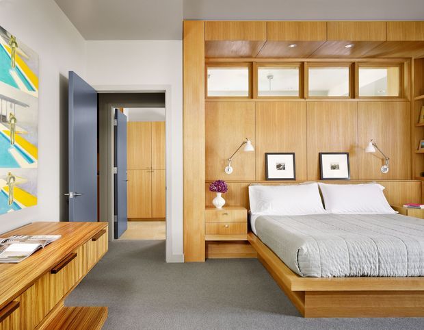 A contemporary bedroom with a built-in timber wall and integrated platform bed. The warm wood joinery includes overhead cupboards, open display shelving, bedside drawers, wall-mounted reading lights, and clerestory-style interior windows above the bed. A grey upholstered bedspread, white bedding, grey carpet, framed artwork, and a timber desk or console complete the room. An open doorway leads to a timber-lined hall or wardrobe area.
