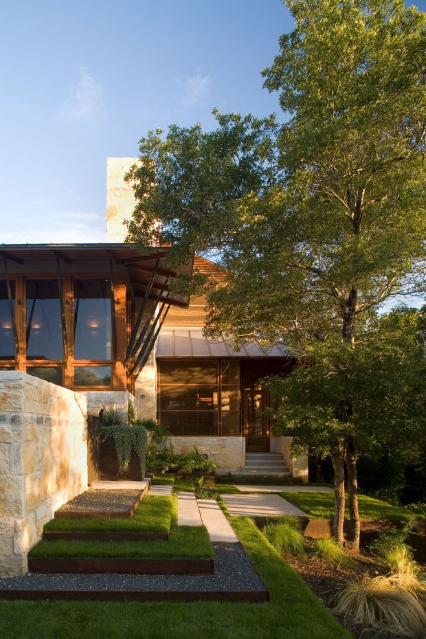 Exterior view of a contemporary multi-level residence set within a landscaped garden. The house features broad glazed openings with dark timber framing, projecting low-pitched roof planes, exposed structural supports, warm timber cladding, and pale masonry or stone walls. A stepped path, gravel strips, clipped lawn terraces, and ornamental grasses lead toward the entrance, with mature trees partially screening the building.