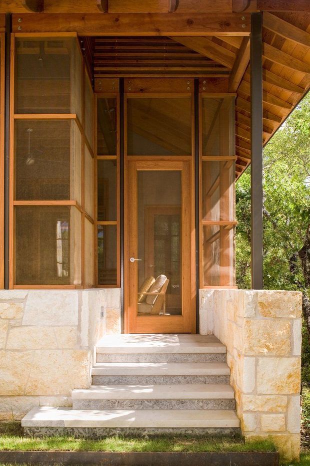 Exterior view of a compact timber-clad entry porch with a glazed timber front door, screened or glazed side panels, exposed timber rafters and a dark slender support post. Pale stone masonry forms the lower walls, and broad pale stone steps lead up from a small lawn. The structure is set among leafy trees and appears to be a carefully detailed small-scale residential or retreat-style entrance.