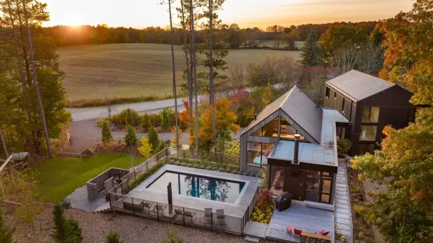 Aerial view of a contemporary multi-level rural residence surrounded by mature trees and open fields. The home features steep gable and pitched metal roofs, dark and light exterior cladding, extensive glazing, terraces, and a rectangular swimming pool enclosed by glass balustrades.