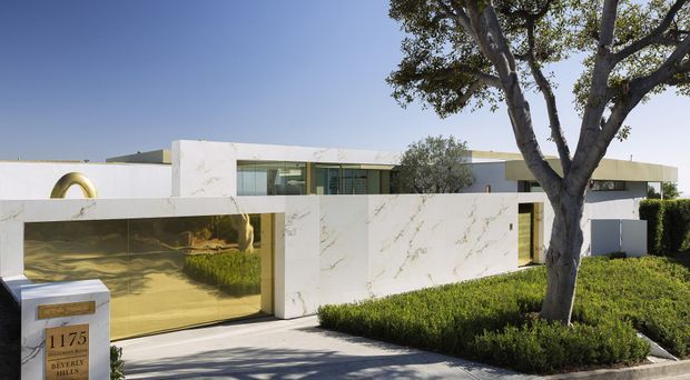 A low-profile, single-storey modern residence viewed from the street. The facade combines white rendered walls with large marble-look panels, extensive horizontal glazing, and a highly reflective gold-toned garage or entry door. Carefully clipped hedges, a mature tree, and a broad paved driveway frame the minimalist frontage. A street number pillar is visible at the boundary.