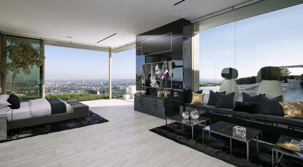 A high-end contemporary living room with floor-to-ceiling glazing, expansive city and water views, pale large-format flooring, a black built-in media wall, and dark modern sofas arranged around glass coffee tables. The open, bright space has strong indoor-outdoor visual connection and refined minimalist furnishings.