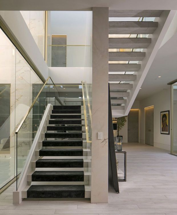 A sophisticated contemporary interior centred on a sculptural open-riser staircase. The staircase has dark stone or marble treads, glass balustrades, and slim brass-toned handrails, rising beside a tall glazed void. Pale stone or marble wall surfaces, light timber-look flooring, concealed warm lighting, and a gallery-like hallway create a refined, spacious composition. A second flight or overhead stair structure crosses the upper right, with framed artwork and minimalist doors visible in the background.