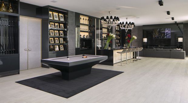 A spacious, sophisticated open-plan entertainment room featuring a black billiards table on a dark rug, extensive dark built-in display and bar cabinetry, a marble-topped island with bar stools, and a lounge area with a large sectional sofa and wall-mounted television. Dramatic chandeliers and integrated lighting contrast with the pale timber-look floor and light ceiling.