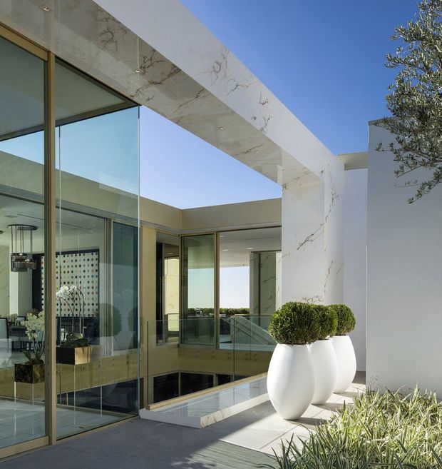 A luxurious contemporary residence viewed from an outdoor terrace or courtyard, featuring floor-to-ceiling glass walls, bronze-toned window and door frames, a dramatic white marble-clad rectangular portal, and sculptural white planters with clipped topiary. Interior dining and living areas are visible through the glazing, with a bright blue sky and carefully landscaped planting completing the scene.