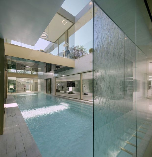 A dramatic multi-level indoor courtyard centered around a long swimming pool, with extensive floor-to-ceiling glazing, a bridge-like upper-level walkway, skylights, and a textured translucent feature wall. The space has a highly modern, open architectural composition with pale stone or concrete surfaces, reflective glass, and cool blue-green water.