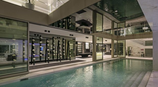 Interior view of a sophisticated indoor swimming pool within a large, multi-level commercial or hospitality space. The pool is surrounded by glass walls, reflective glazing, dark framed doors, illuminated display or lounge areas, and an upper-level balcony. The space has a polished, contemporary architectural character with dramatic reflections and subdued lighting.