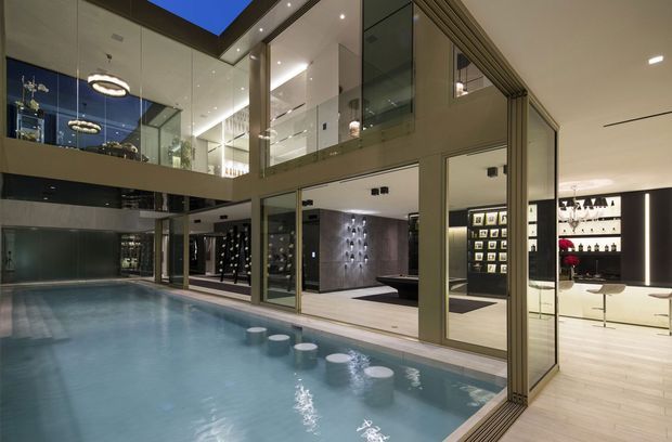 A sophisticated multi-level interior centered around a large indoor swimming pool, with extensive glass walls and doors opening to a bright gallery-like living and entertaining space. The space includes a dark stone feature wall, display shelving, framed artwork, a bar or drinks display area, recessed ceiling lighting, and an upper-level mezzanine or balcony. The overall design is luxurious, contemporary, and strongly connected through transparent glazing.