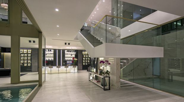 A spacious, high-end commercial interior, likely a retail showroom or reception area, with a polished light floor, illuminated wall displays, extensive glass partitions, and a prominent contemporary staircase. The staircase has a white sculptural underside, glass balustrades, and slim gold-toned handrails. A large mirror on the left reflects the display area, while a glass-enclosed stair and upper-level void create an open, multi-level composition.