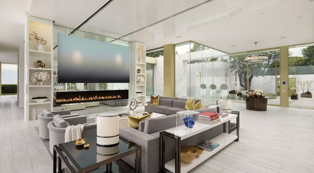 A bright, spacious contemporary living room with floor-to-ceiling glazing, a large wall-mounted television, linear fireplace, grey upholstered sofas and armchairs, and open white display shelving. The room has pale timber-look flooring, glossy white ceilings, a glass feature wall, and restrained metallic and neutral furnishings. A landscaped outdoor area is visible through the glazing.