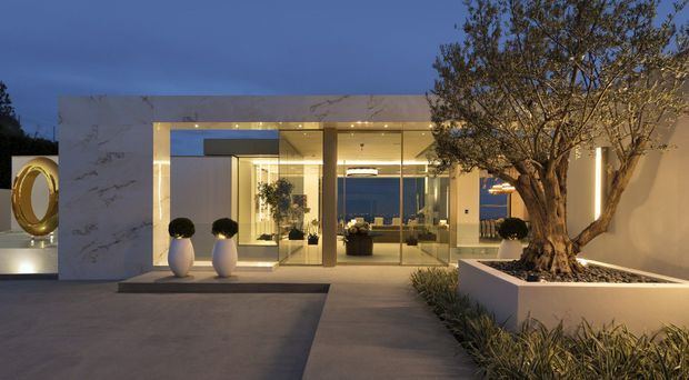 Contemporary luxury residence exterior at dusk, featuring a broad pale stone or marble-clad entry portal, floor-to-ceiling glazing, warm recessed lighting, a refined paved forecourt, and a raised planter containing a mature sculptural tree. The transparent entry reveals a softly lit interior and distant city or landscape views. A large circular metallic feature is visible at the far left.
