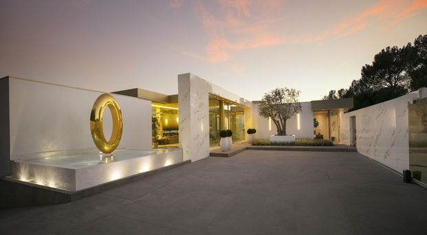 A contemporary low-rise residence photographed at dusk, centred on a broad paved forecourt and courtyard. The building has crisp white rendered volumes, large glazed openings, a flat roofline, illuminated interior spaces, and a prominent circular metallic sculpture set on a raised reflecting or display plinth. A white marble-look wall and landscaped planting frame the entrance, with mature trees visible behind the property.