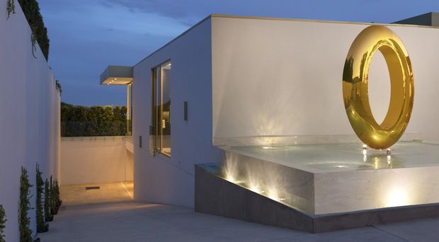 Twilight view of a minimalist modern architectural exterior, featuring crisp white rendered walls, a flat roof, narrow vertical glazing, recessed architectural lighting, and a raised pale stone or concrete plinth. A large polished gold circular sculpture forms the main feature, while a narrow landscaped passage leads alongside the building.