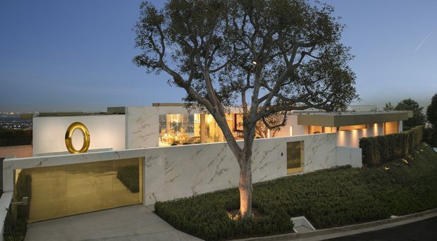 Twilight exterior view of a low-slung contemporary residence on a landscaped, sloping site. The home has crisp white rendered walls, prominent marble-look wall panels, extensive warm-lit glazing, a flat roofline, a sculptural circular gold feature on the facade, and a concealed garage integrated into the front elevation. A mature tree and manicured hedging frame the composition.