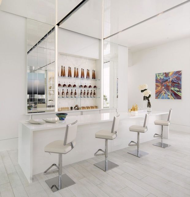 Bright, highly contemporary white home bar with a long glossy island and four modern chrome pedestal bar stools. Mirrored cabinetry and shelving display bottles and glassware, while white walls, pale timber-look flooring, a colourful abstract artwork, and a floral arrangement complete the refined entertaining space.