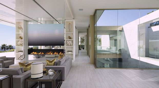 Bright contemporary living room with a large sectional sofa, coffee tables, built-in shelving, a wall-mounted television, and a linear fireplace. Pale timber-look flooring and white built-in joinery create a light, refined interior, while extensive floor-to-ceiling glazing opens views toward a modern courtyard and adjacent spaces. A corridor extends beyond the living area.