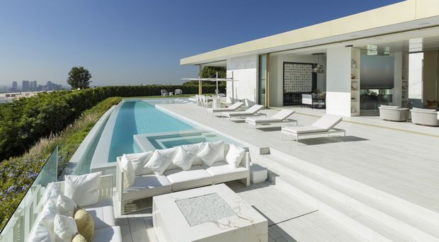 A bright, high-end modern rooftop or elevated outdoor terrace with a long rectangular swimming pool, pale tiled decking, white outdoor sofas and sun loungers, and a contemporary flat-roofed white pavilion opening onto the pool area. The space overlooks a distant urban skyline and is bordered by manicured hedging and planting.