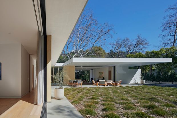 View from a contemporary interior through full-height glazed sliding doors toward a low, flat-roofed pavilion and landscaped courtyard. The building features crisp white rendered walls, extensive glazing, timber-clad structural elements, broad roof overhangs, and an open-plan outdoor seating area. Mature trees and lawn surround the architecture under clear daylight.