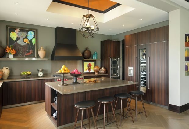 A sophisticated contemporary kitchen with a large central island, dark timber cabinetry, a pale stone benchtop, integrated stainless-steel appliances, and four bar stools. The room features a dramatic black rangehood, dark green-grey walls, warm timber flooring, a geometric pendant light, and a recessed ceiling detail with warm accent lighting. Artwork and decorative objects add colour and personality.