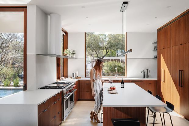 Bright, contemporary kitchen with a large white island, integrated seating, timber cabinetry, stainless-steel appliances, and expansive timber-framed windows overlooking greenery and a pool. The space has a clean, refined feel with abundant natural light and a warm timber-and-white palette.
