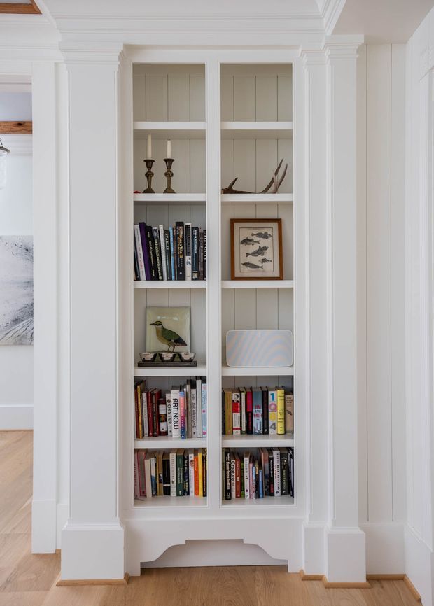 A tall, built-in white bookcase with two narrow sections is recessed into a traditionally detailed wall. The shelving contains books, candlesticks, framed artwork, decorative objects, and a small speaker. Surrounding architectural details include painted wall panelling, substantial classical mouldings, pilasters, a deep cornice, and stepped skirting boards. Light timber flooring and a bright neutral palette create a refined, quietly traditional interior.