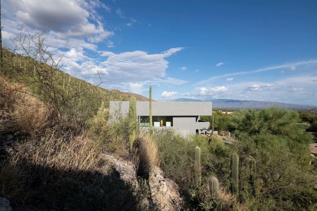 A modern, rectilinear flat-roofed house set into a dry, rugged desert landscape. The pale concrete or stucco-like facade has crisp horizontal and vertical volumes with dark recessed glazing, surrounded by native scrub, tall cacti, rocky terrain, and distant mountains under a bright blue sky.