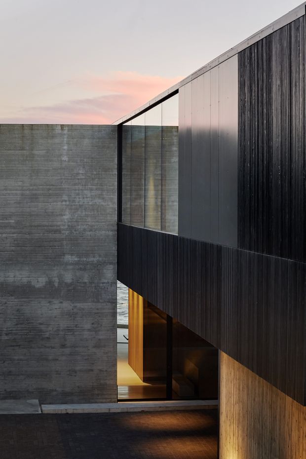 A dramatic exterior architectural view of a contemporary multi-level building, featuring a tall raw concrete wall, extensive dark vertically ribbed cladding, slim dark window frames, and large glazed openings. Warm timber surfaces and interior lighting are visible beneath a projecting upper volume, with a paved entry area in the foreground and a soft dusk sky beyond.