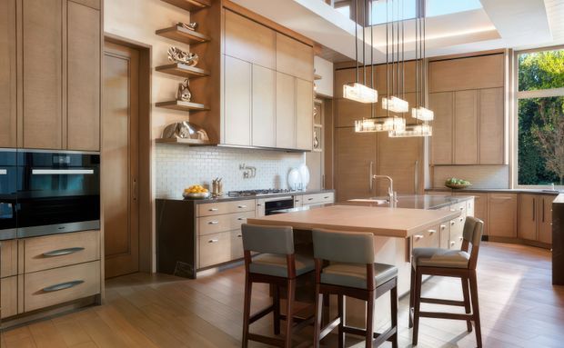 A spacious, refined kitchen with extensive warm timber cabinetry, integrated black appliances, a large central island with a timber benchtop, and seating for four. The kitchen includes a white small-format tiled splashback, open display shelving, a large window, clerestory glazing, and a cluster of contemporary pendant lights over the island. The overall design is warm, bright, and sophisticated.