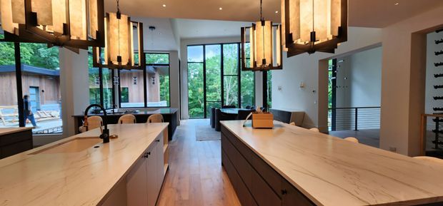 A spacious contemporary kitchen viewed from between two large stone-topped islands. The room features extensive light-toned benchtops, dark wood cabinetry, warm timber flooring, black-framed floor-to-ceiling windows, and multiple oversized rectangular pendant lights. An open living or dining area is visible beyond the kitchen, with a wine-storage wall at right and lush greenery visible through the glazing.