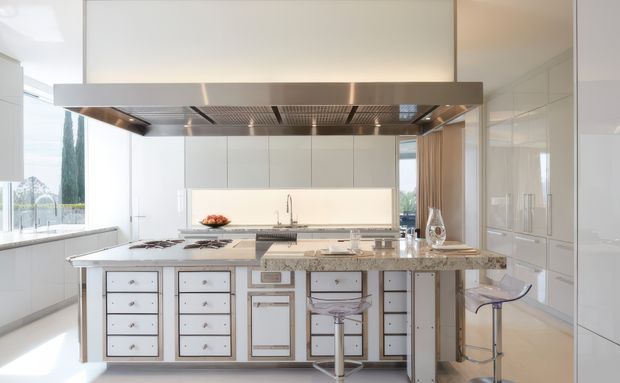Bright, spacious contemporary white kitchen with a large central island, integrated cooktop and expansive stainless-steel range hood. The island features white drawer cabinetry with metallic trim, a pale stone benchtop, undermount sink, and clear bar stools. Glossy white full-height cabinetry, a light splashback, and large windows create an airy, polished interior.