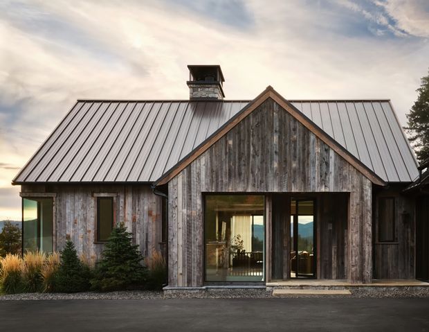Exterior view of a rustic single-storey gabled home with weathered vertical timber cladding, a standing-seam metal roof, and a projecting timber-framed entry volume. Glazed doors reveal a glimpse of the interior, while landscaped planting and a paved driveway sit in front of the building.