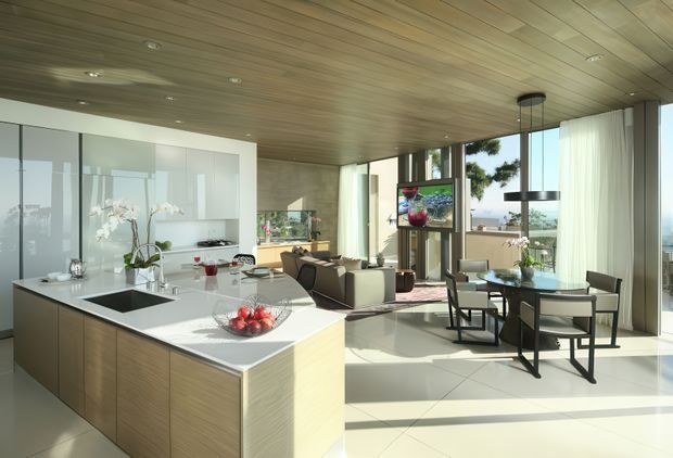 A bright open-plan contemporary kitchen and dining/living area with a large timber-panelled island, white stone benchtop, integrated cooktop and sink, and sleek white cabinetry. The room features a warm timber-lined ceiling, extensive floor-to-ceiling glazing, pale tiled flooring, minimalist furnishings, and views toward the outdoors. A dining table and seating area sit beside the kitchen, with a pendant light above.