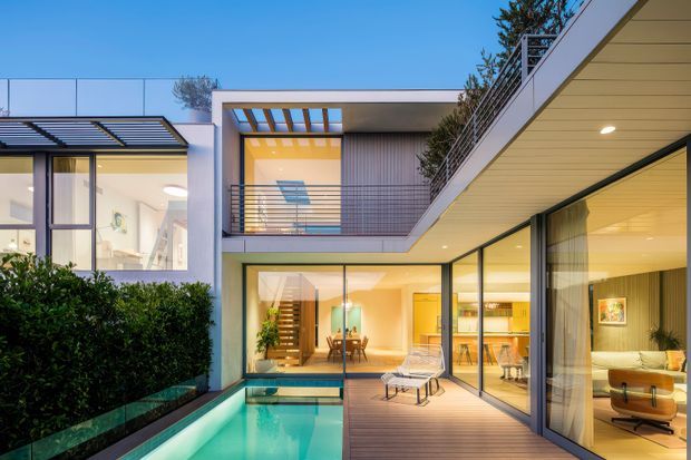 Contemporary multi-level residence arranged around a narrow turquoise swimming pool and timber deck courtyard. The image shows extensive floor-to-ceiling glazing, slim dark window frames, clean white rendered forms, a sheltered outdoor living area, an upper-level balcony, and warm illuminated interiors visible through the glass. A pitched pergola and vertical screening add architectural detail.